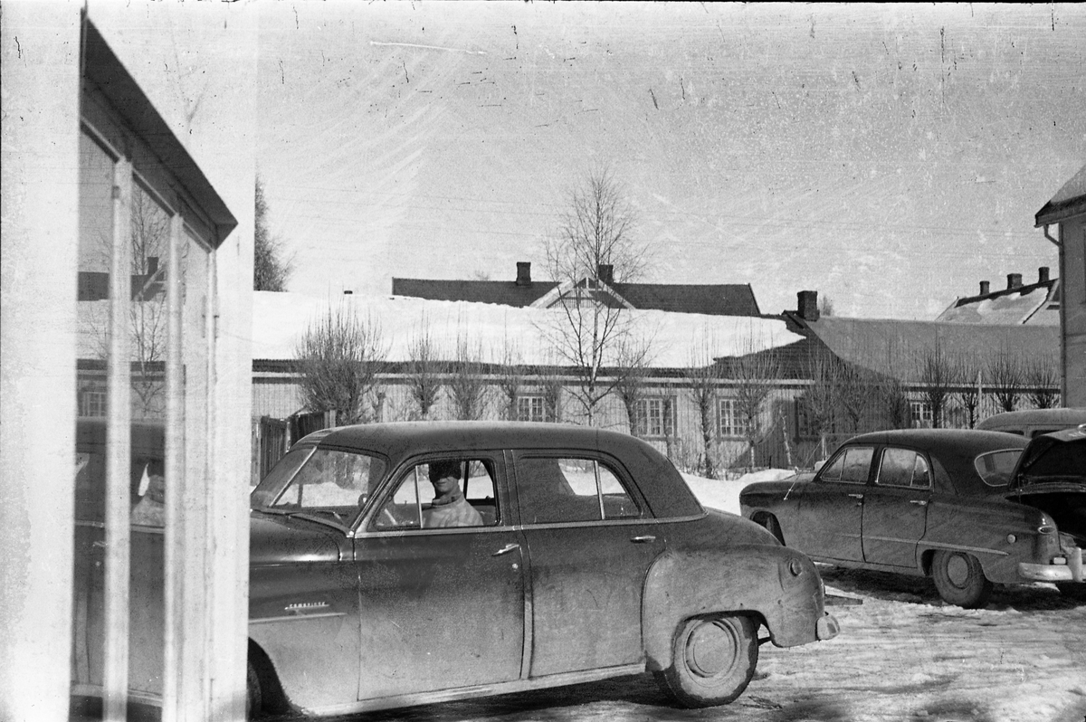 Fra Lena sentrum mars 1954. Mauritz Enger sitter i bilen, som må antas å være hans drosjebil, ved drosjebua ved Esso stasjonen. Bilen Mauritz sitter i er iflg. informant en Dodge eller Plymouth  av årsmodell 1948-52. Et annet bilde tatt ved en annen anledning viser imidlertid at det er en Plynouth, årsmodell 1951-52).  Bilen bak, med åpent bagasjelokk er iflg. samme informant en amerikansk Ford 1949-51.