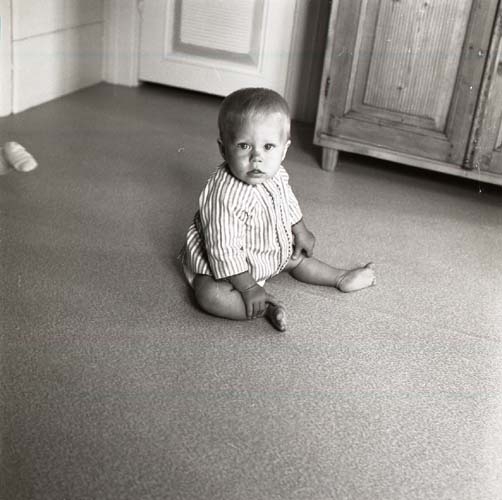 Ett barn sitter iförd en skjorta på golvet. Intill syns benen på ett golvskåp, 1971.
