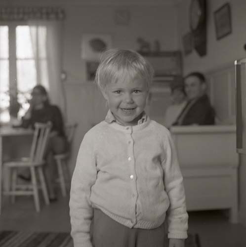 Ett barn poserar för kameran i förgrunden. I den disiga bakgrunden skymtar tre vuxna personer fram, 1968.