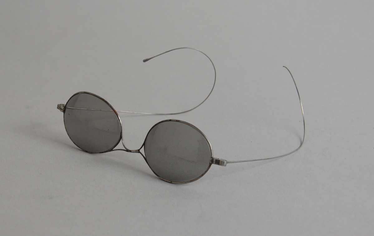 Solbriller med ovale mørke glass og metallinnfatning, med tynne metallbøyler.
