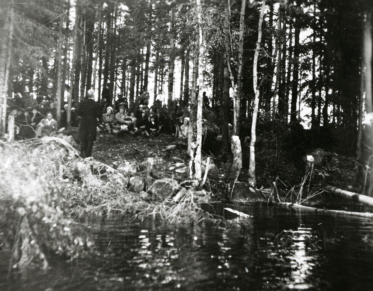 En man talar inför en grupp människor i en skogsglänta intill vatten.