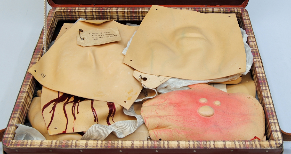 En utbildningsväska av handkofferttyp, gjord av hårdpapp och lackad i roströd färg. På väskans sidor sitter läderförstärkningar i form av infärgat läder fastsatta med metallnitar. Väskan har axelband av brunt läder samt två låsbara lås av metall. Två nycklar i rött band hänger på axelbandet, på bandet återfinns en "metallsigill" med märkningen "Aktiebolaget Tidan Unica Mariestad".
Väskan är inredd med en träram klädd i rutigt papper. Den övre inredningen består av två fackdelar av trä, även det i roströd färg. De övre facken består av sex olika fack, den nedre av två större. Facken är märkta med siffror i gult tryck.
Väskan har även två spännband för att innehållet ska hållas på plats.

Väskan innehåller 25 stycken sårattrapper av gummi som imiterar diverse skador. 
1 för ansikte (:1)
1 för öra (:2)
1 för hand (:3)
2 för fot (:4-5)
20 stycken för oidentifierbara delar av kroppen. (:6-25)
1 kuvert med innehåll (:26)
1 väska (:27) 
6 lappar med beskrivning av de olika skadorna finns för att fästa på attrapperna. Varje attrapp har en märkning i form av nummer som sätts ihop med lapparna. En av attrapperna har tillverkarnamn tryckt på baksidan. Det finns också gummiband, resårband och metallhyskor för fästning av attrapperna i lådan, liggandes i ett kuvert märkt med tillverkarnamn. Handledningsmanual från Röda Korset återfinns det med.