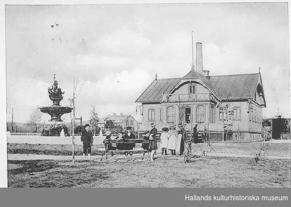 Vykort daterat 1902-11-22 föreställande varmbadhuset i Kungsbacka med en stor porlande fontän i den spirande parken. En grupp barn är samlade kring en bänk och två kvinnor står med handdukar - kanske personal på badhuset. Ur bild ligger järnvägsstationen till vänster.