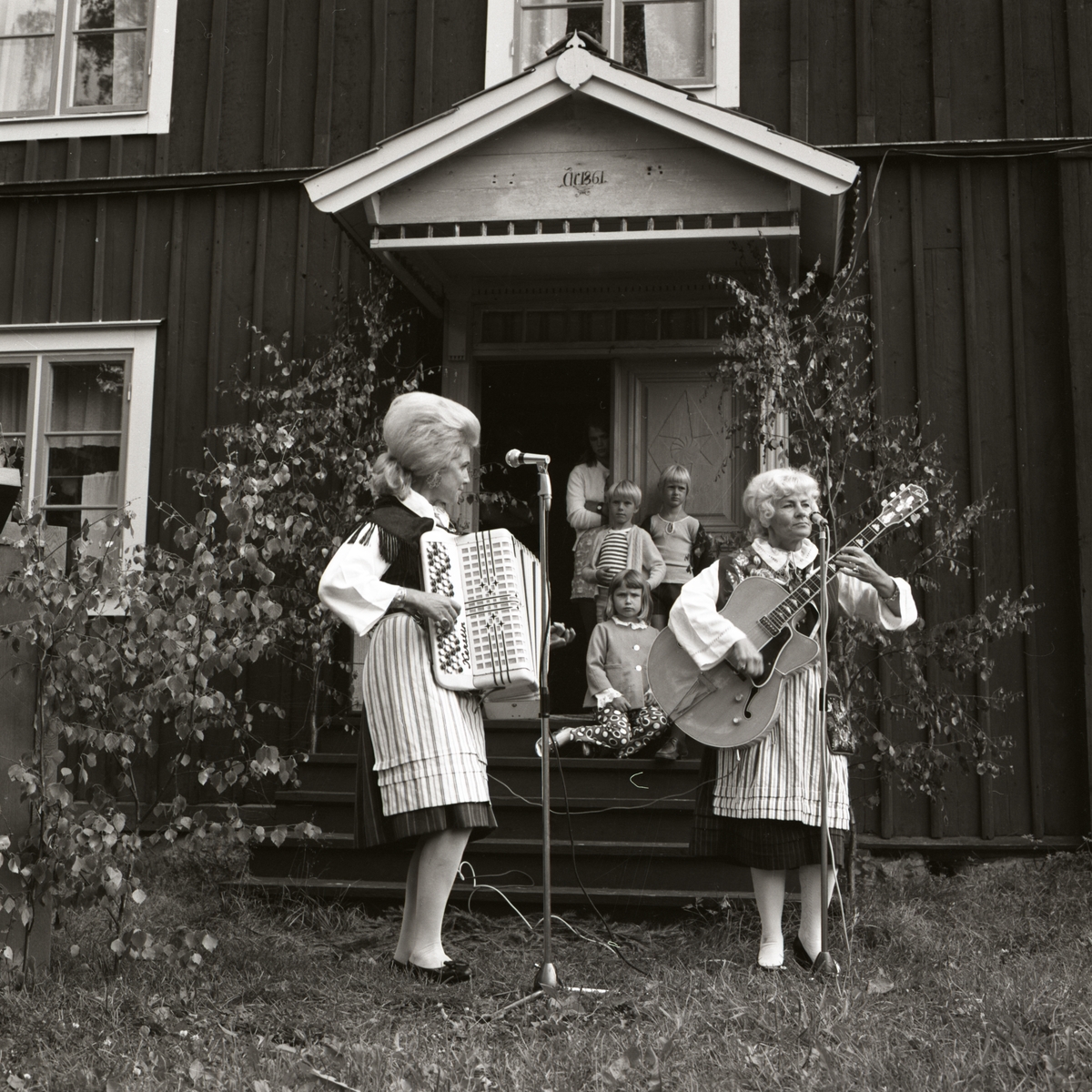 Två kvinnor underhåller vid Rengsjöfesten på hembygdsgården. Några barn står under brotaket och lyssnar på kvinnorna som spelar dragspel och gitarr, 1970.