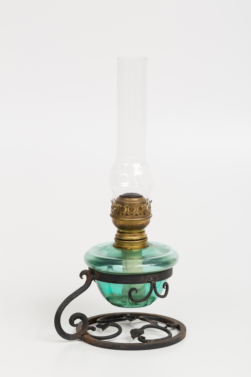 Parafinlampe i 3 deler; parafinlampe (, stativ til lampe og lampeglass. Lampeglasset er blitt knust og fått ett hull. Smidd dekorasjon på stativet.