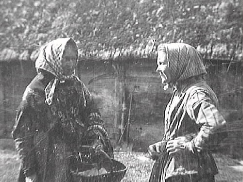 Charlotta, Severins syster, samtalar med en kvinna ute på gårdsplan. Charlotta står med handen i sidan (till höger). I bakgrunden syns en lada med stråtak. Björsgård.