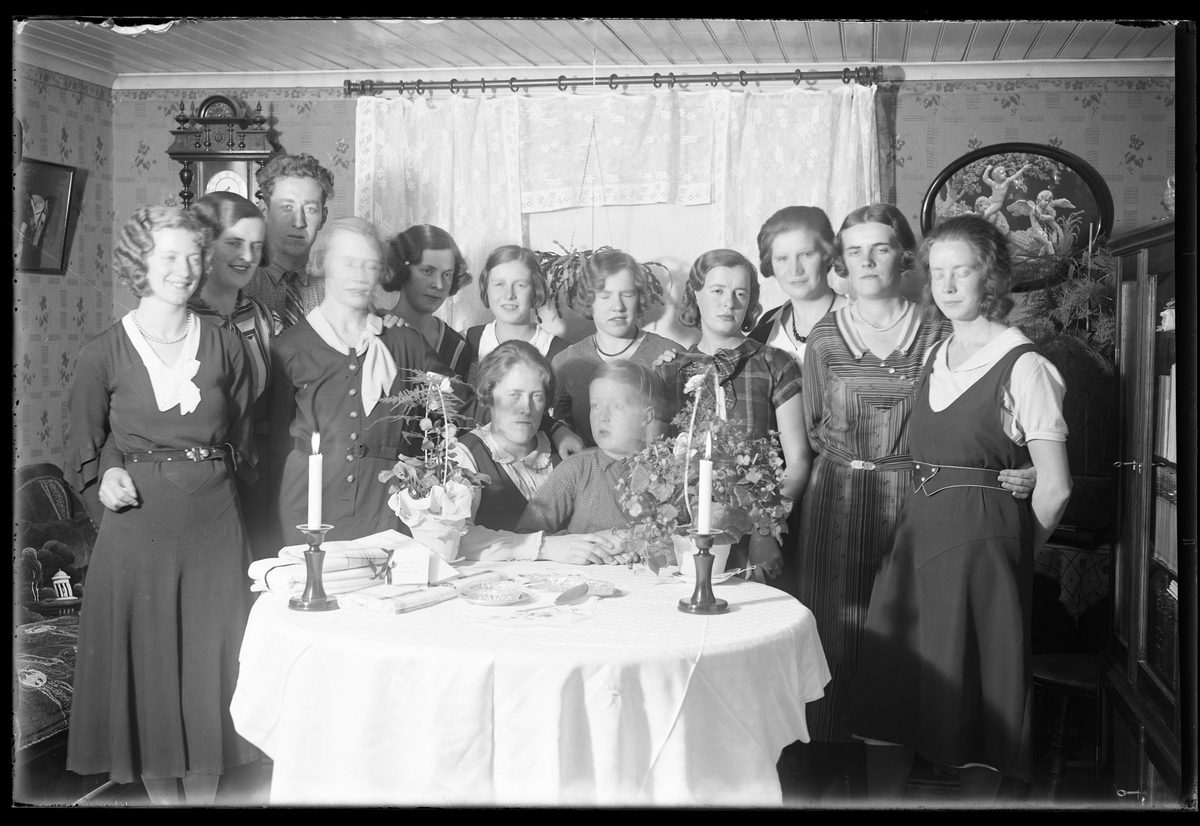 Elva kvinnor, en man och en pojke är samlade runt ett bord. På bordet ligger en vit duk och två ljus är tända. I fotografens anteckningar står det "Antonssons, Vadsjön".