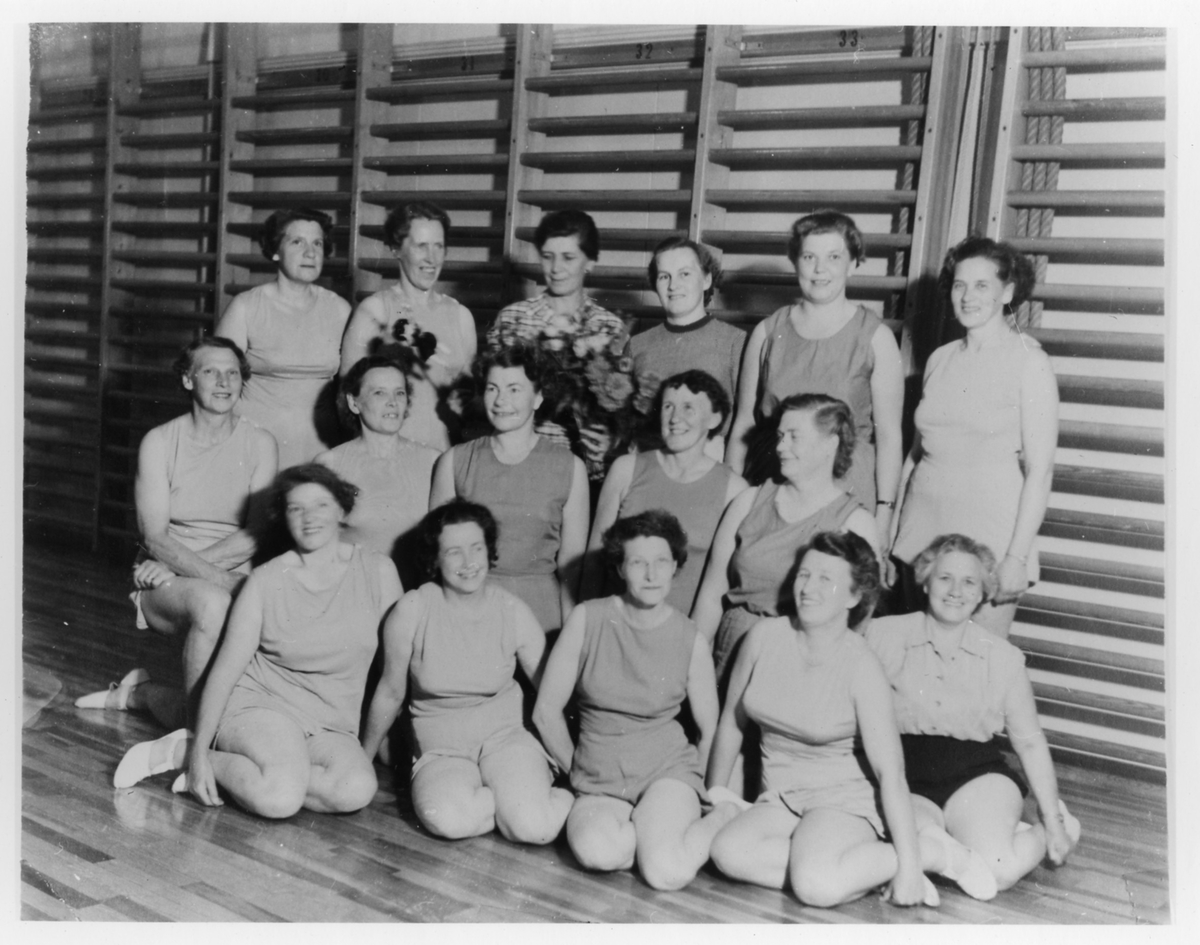 Gruppbild av 16 gymnastikklädda kvinnor, stående och sittande i en gymnastiksal, från Alingsås Kooperativa Kvinnogille. Kvinna i bakre raden håller en bukett blommor.