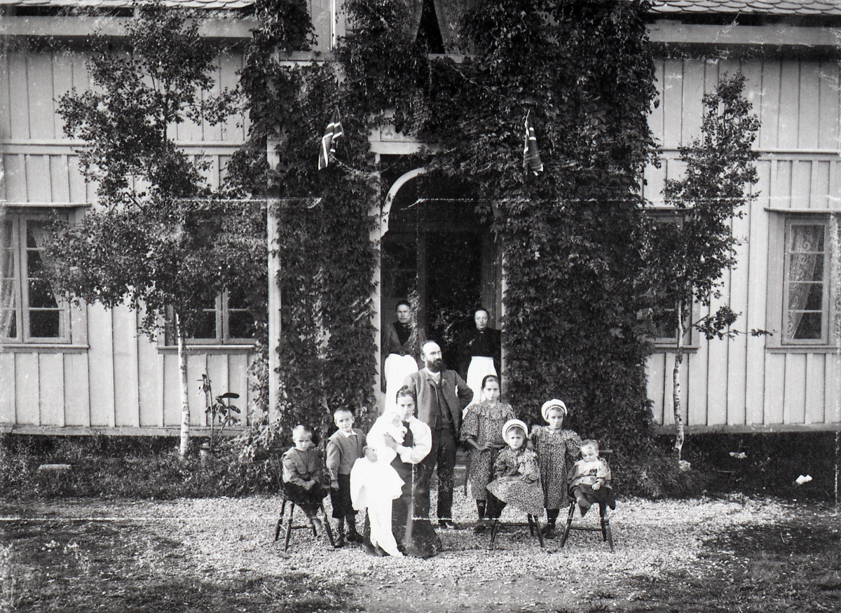 Sokneprest J.C. Sørlie med kona og 7 barn utenfor prestegården. I døra står to kvinner. Den eine er Guri Haugen.