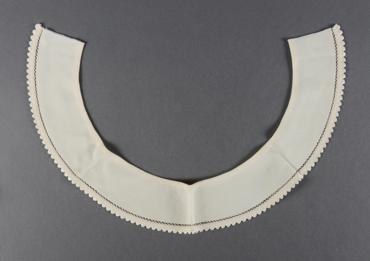 En buet løskrage laget av hvitt kunststoff. Langs den øvre/indre kanten er stoffet foldet inn. Langs den ytre kanten er det sydd en enkel bord og sydd på et hvitt tungebånd.