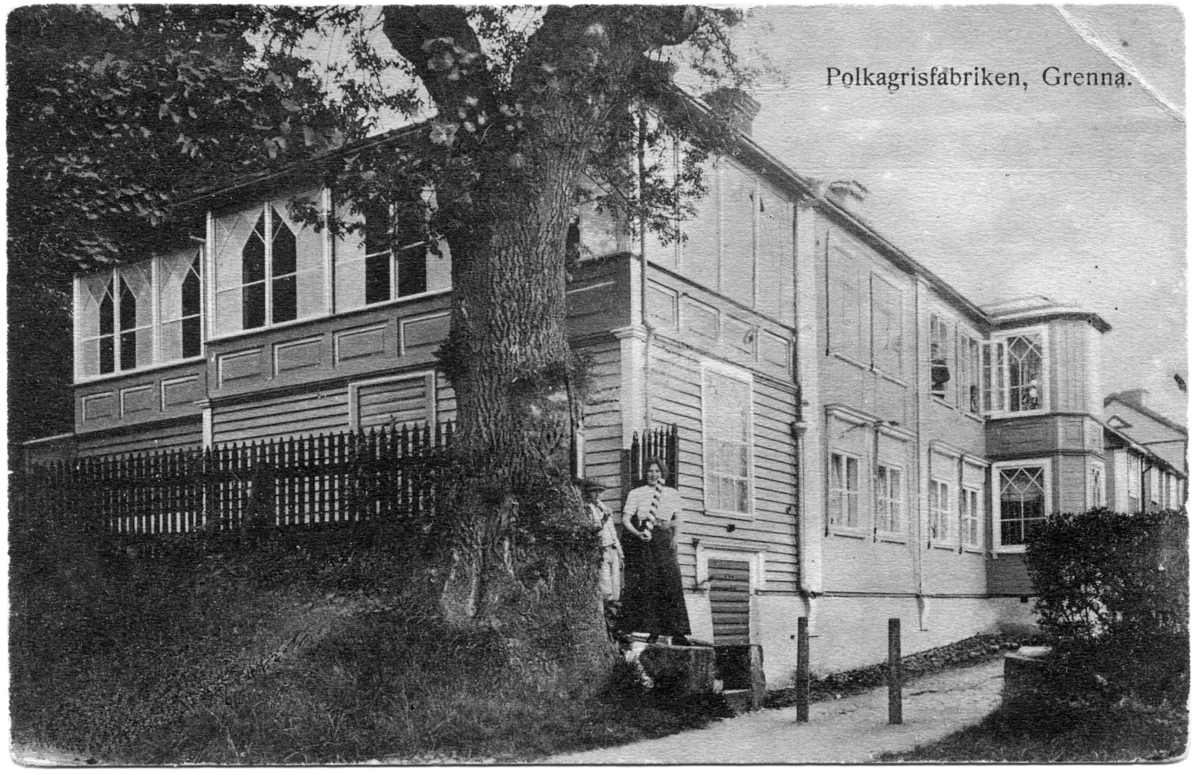 Fastigheten Bergsgatan 14 sedd från Gränna kyrka. I hörnet står en flicka och en pojke. Hon har en stor polkagris i handen och han äter på en polkagris. I fastigheten bodde polkagristillverkaren Amalia Erikson åren 1911-1915.