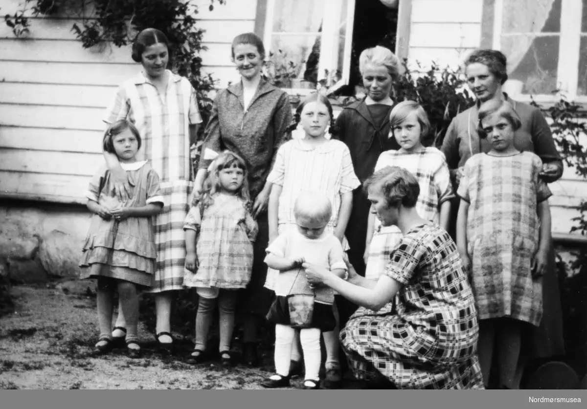 Foto av en gruppe kvinner og barn, oppstillt utenfor et bolighus. Fra Fru Williams fotoalbum. Eier av originalmaterialet er Gunnar Williams. Fra Nordmøre museums fotosamlinger.