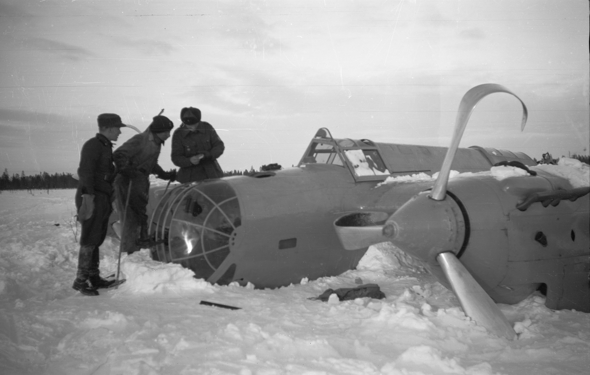 Nedskjutet sovjetiskt bombflygplan SB-2 ligger på ett fält vid Svenska frivilligkåren i Finland, F 19. Tre militärer undersöker nospartiet.