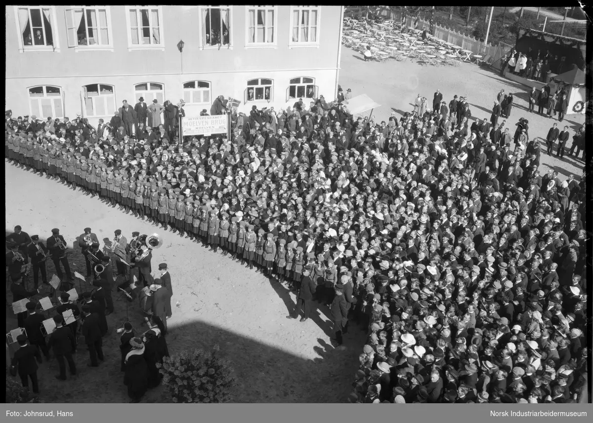 Åpning av Fylkesutstillingen 1922 med besøk av Kong Haakon VII. HM kongen står på rød løper. Korps spiller, folkemengde rundt hovedarena for åpningsseremonien.
