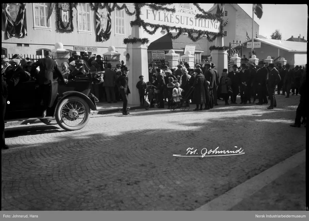 Fylkesutstillingen 1922. Mennesker i gaten og sittende i parkert bil foran inngangen til utstillingen.