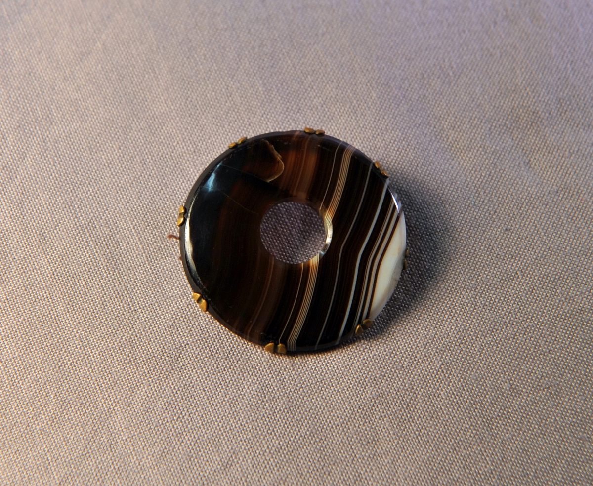 Beskrivning: En st. rund brosch i form av en brun-vitrandig agat med hål i mitten. Infattad i mässing.