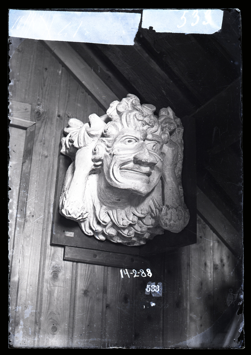 Gipsavstøpning av skulptur fra Nidarosdomen, plassert mellom tårnbuene innvendig. En person med vilt hår og et karikert ansikt som holder seg til håret og ansiktet.