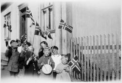 Barn feirer 17. mai med flagg og tromme, ved hus og gjerde.