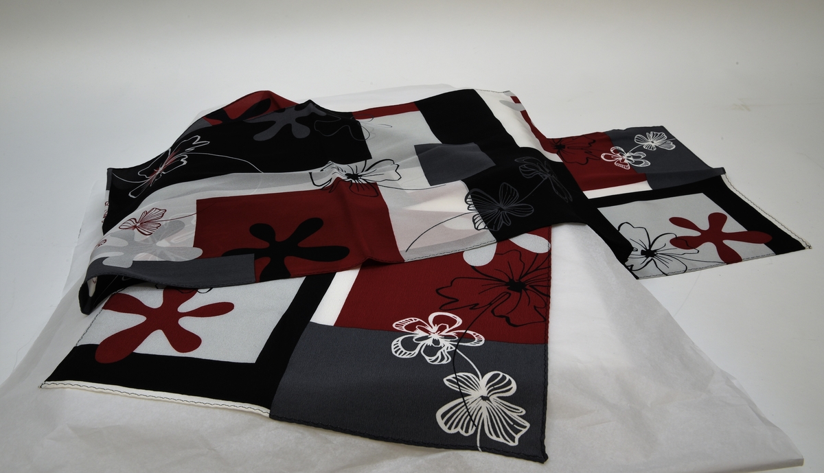 Scarf av silke med ett upprepande mönster bestående av blommor och geometriska figurer i rött, grått, vitt och svart. Scarfen har två lappar fastsydda vid ena kortsidan.