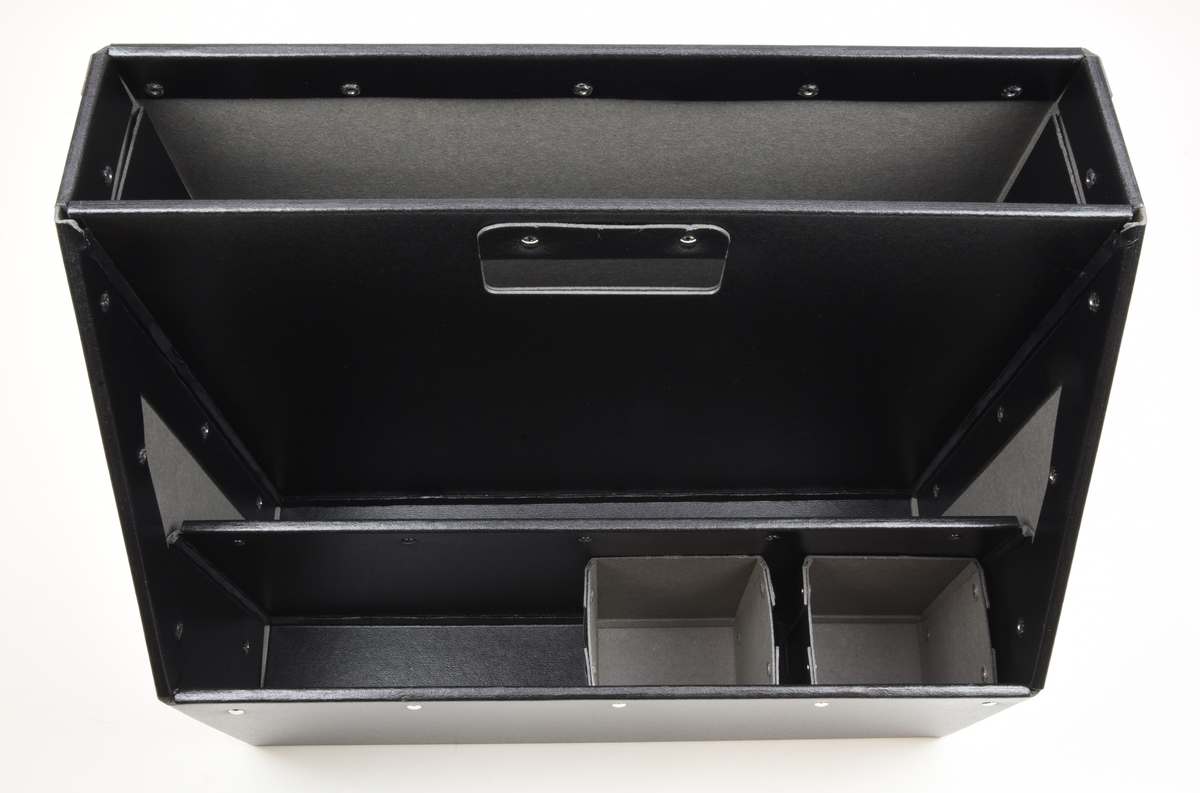 Bärbar förvaringslåda tillverkad av Bigso AB, modell ”Carrie”. Lådan är tillverkad av pressad pappersfiber som nitats samman och har en svart, lätt skrovlig yta. Formen är rektangulär med en avfasad framsida för lättare åtkomst av de tre facken. I den mittersta avdelaren finns ett utstansat handtag. I det främre facket finns två lösa smålådor avsedda för till exempel pennor. På lådans vänstra gavel finns en liten plastficka med ett vitt papper för märkning av lådan.