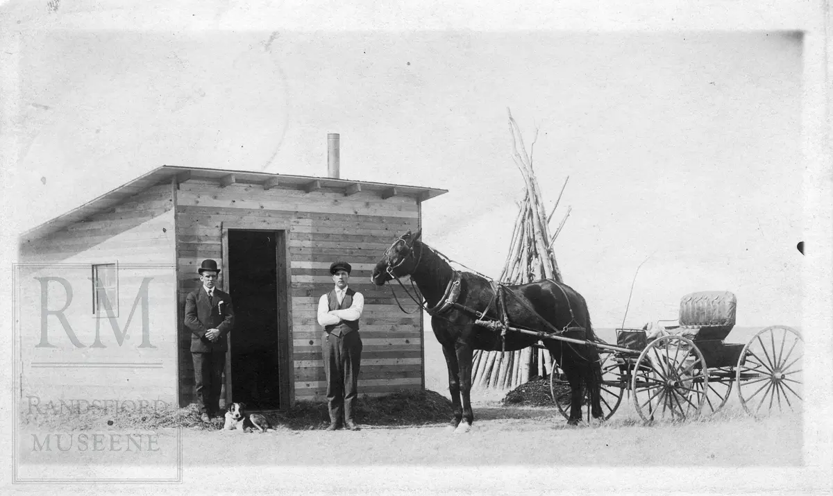 To menn og hest med vogn foran en hytte. En hund ligger ved bena til mannen til venstre. "Fra Kopperuds første homestead."