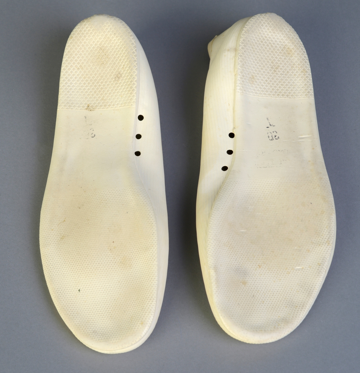 Et par hvite badesko laget av gummi. De har form som en tøysko/tøffel. Det er støpt striper inn i gummien slik at det minner om tekstil. På innsiden av skoen er det tre luftehull. I sålen er det støpt finrutet mønster i hælene. På undersiden av sålen er det støpt rutetmønster på hælen og i front. Det er et svart stempel på undersiden av begge skoene. Størrelsen er 38.