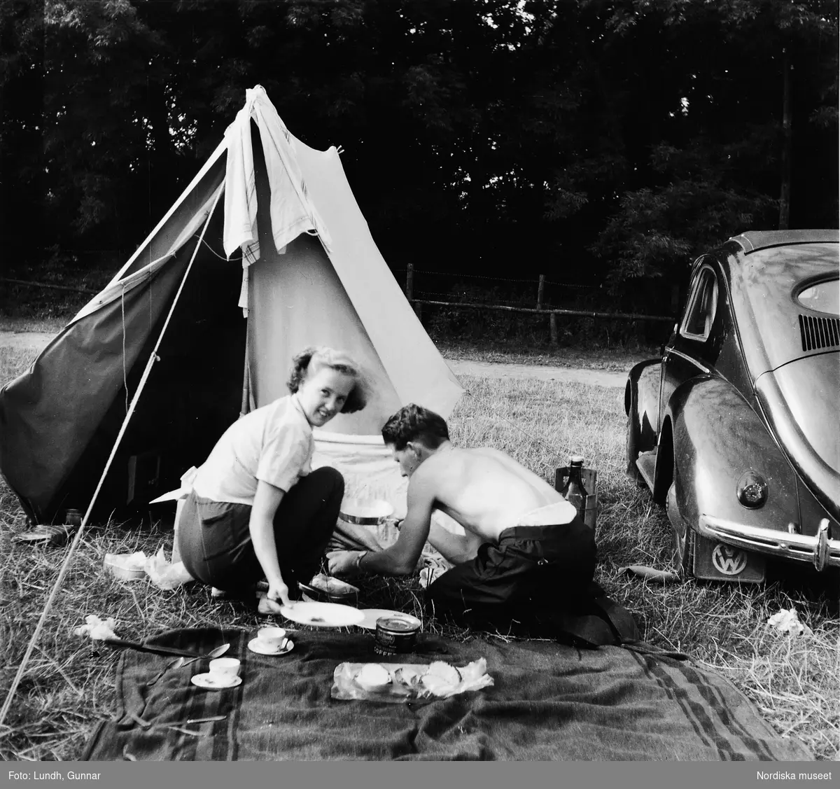 En kvinna och en man sitter utanför ett tält och lagar mat. Bredvid tältet står en bil. Bilden tagen i Skåne år 1955.