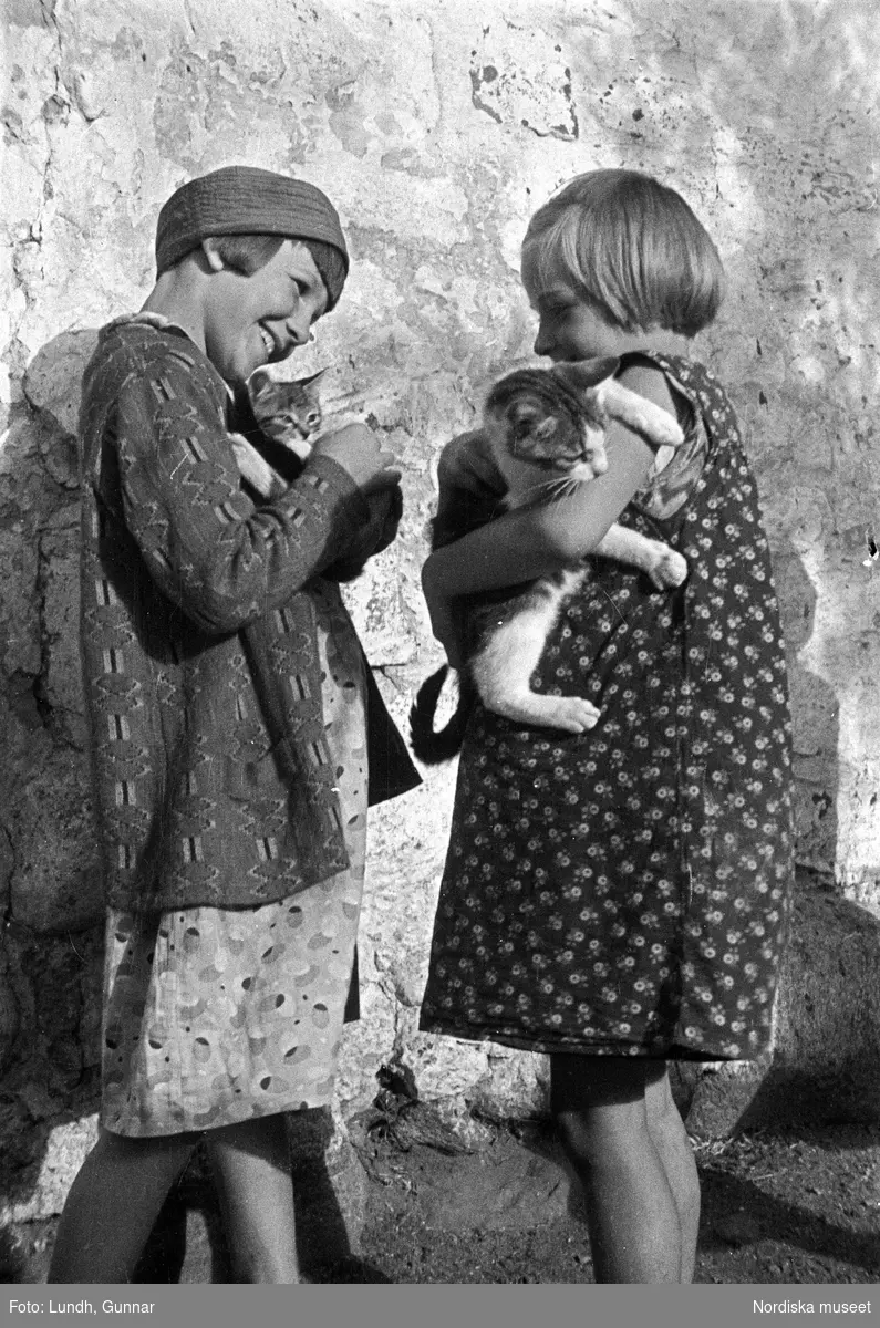 Porträtt av två flickor som står vid en husvägg och håller en katt i famnen, porträtt av en flicka som håller en katt i famnen, fotograferat vid Kullen (Kullaberg), Strandbaden, Tallbacken.