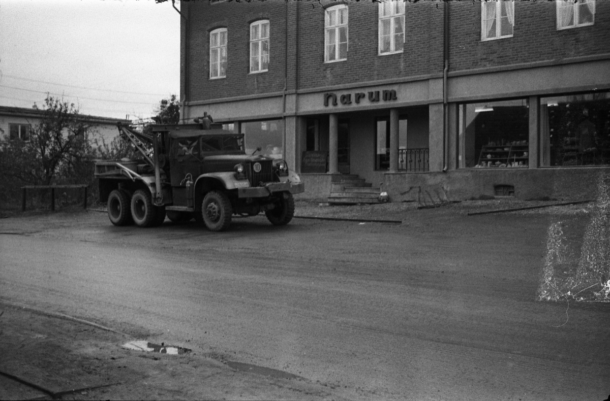 Gatebilde fra Lena sentrum september/oktober 1957. Bilen som står foran Narums-butikken er en Diamond T(969A?) Wrecker (bergingsbil) fra forsvaret. Bildet er tatt gjennom vinduet i Lena Foto & Radio i "Raubua".
