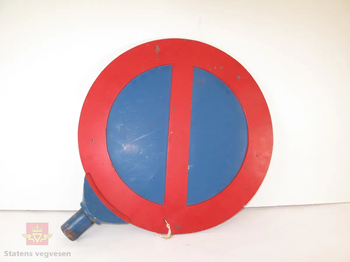 Støpt aluminiumsskilt med holder. Sirkelformet skilt med rød ramme (7 cm) og en rød strek på skrått over skiltet. Det har blå bunn og er et forbudsskilt som angir at parkering er forbudt. Det er to små skruehull på hver side mot nedre del av skiltet. Holderen er malt blå, og ser ut til å være formet for å kunne settes ned i en stolpe eller et rør.
