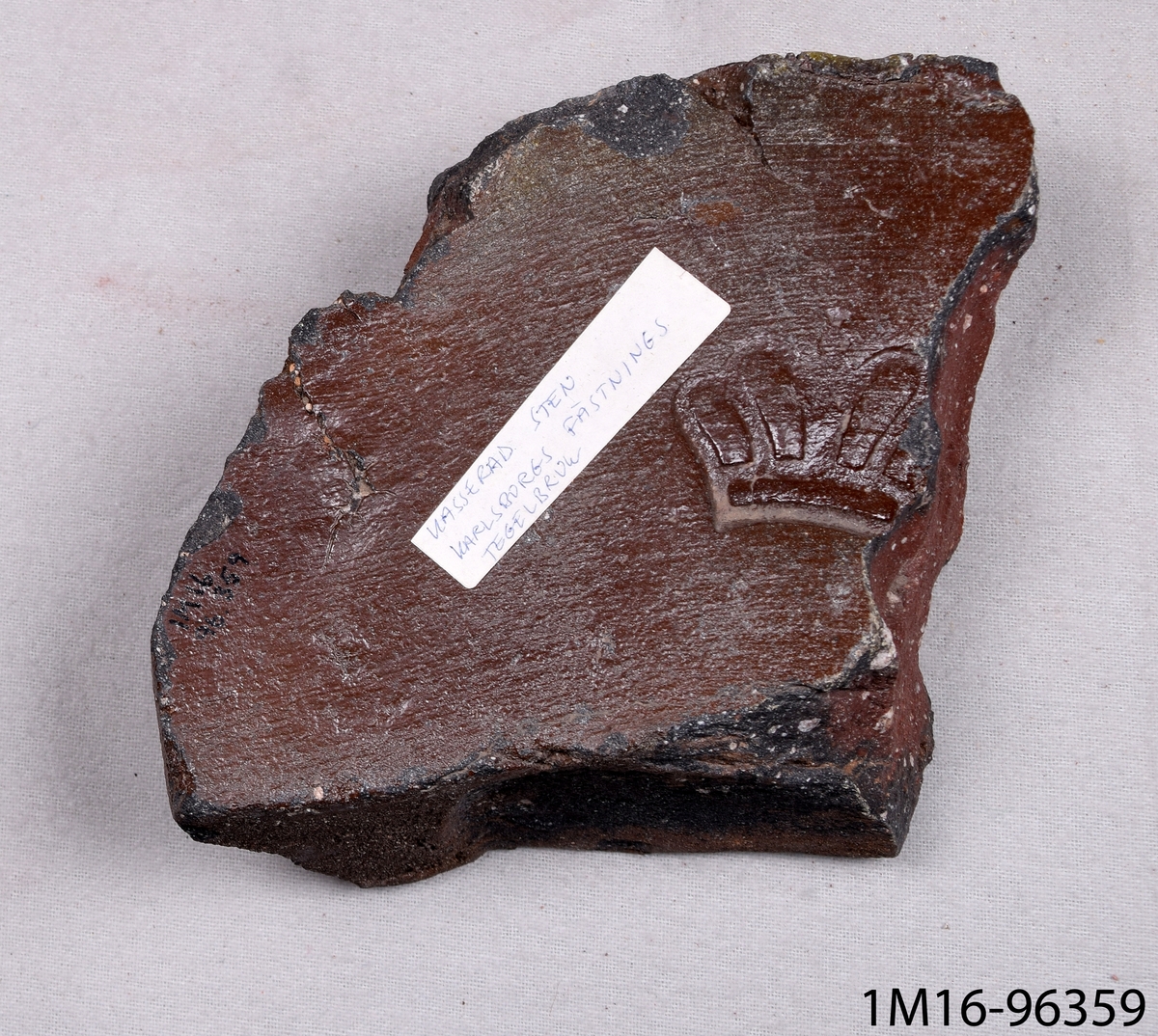 Fragment av murtegel, av bränd lera med brun glasyr. Har ett kronliknande ornament. Från Karlsborgs fästnings tegelbruk.