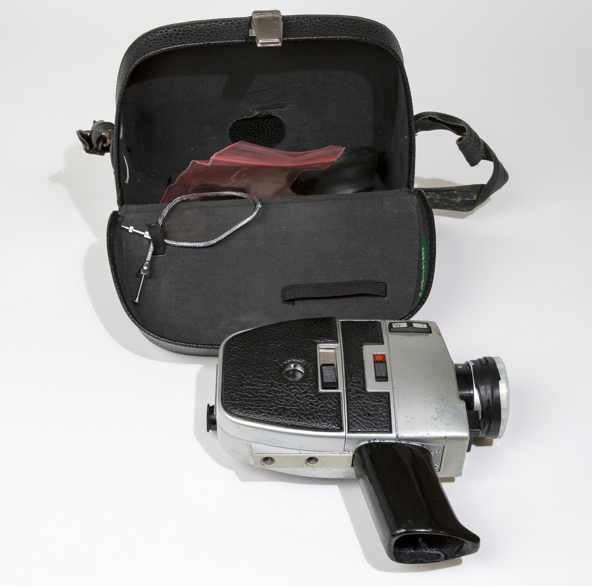 Filmkamera, super-8 BAUER C ROYAL 6-zoom med kamerahus av grå metall delvist klädd med svart skinnimitation. Handtaget är av svart plast och har utrymme för batterier och tillsluts av en lös platta. På linsen sitter ett genomskinligt linsskydd. Filmkameran är placerad i en svart väska, JM 56586, i vilken finns en fjärrutlösare, ljusskydd av gummi för linsen, liten gummiring för sökaren, och större gummiring.