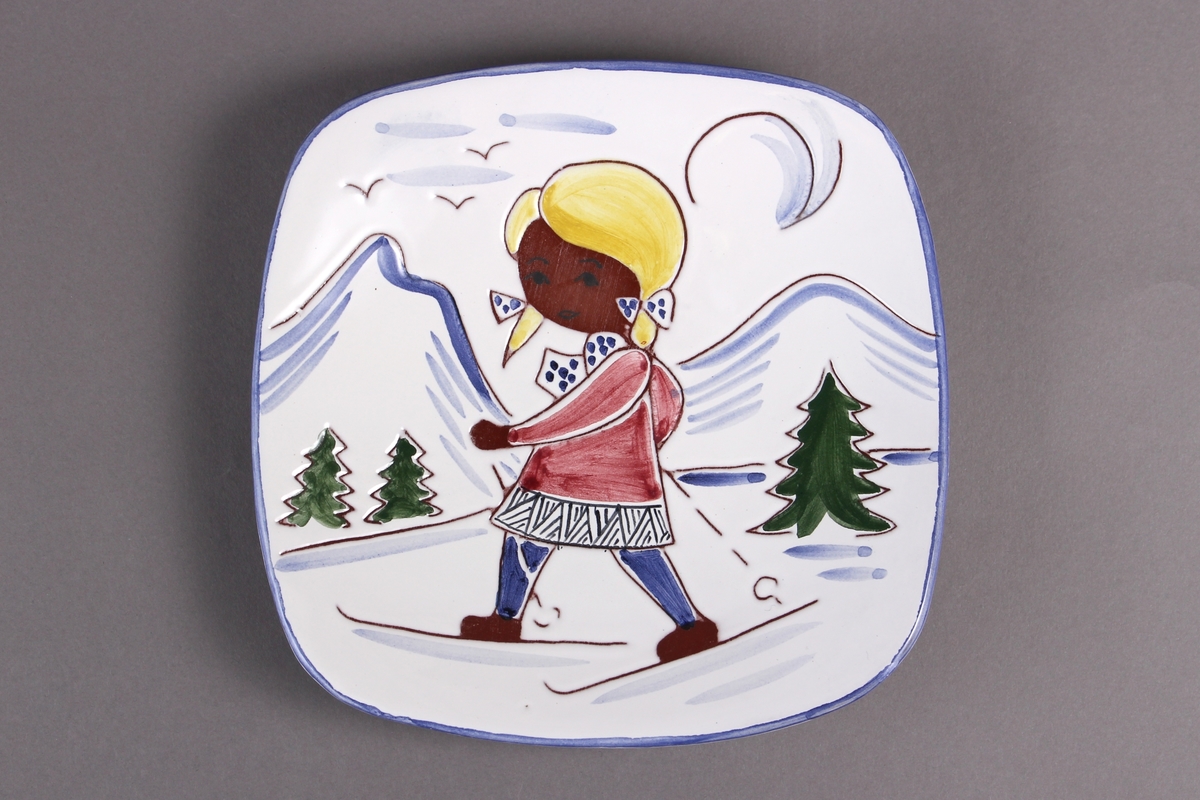 Asjett dekorert med motiv som viser en jente som går på ski i vinterlandskap. Med hull til oppheng på baksiden.