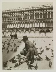 Mann sitter på huk med duer på åpen plass i Venezia