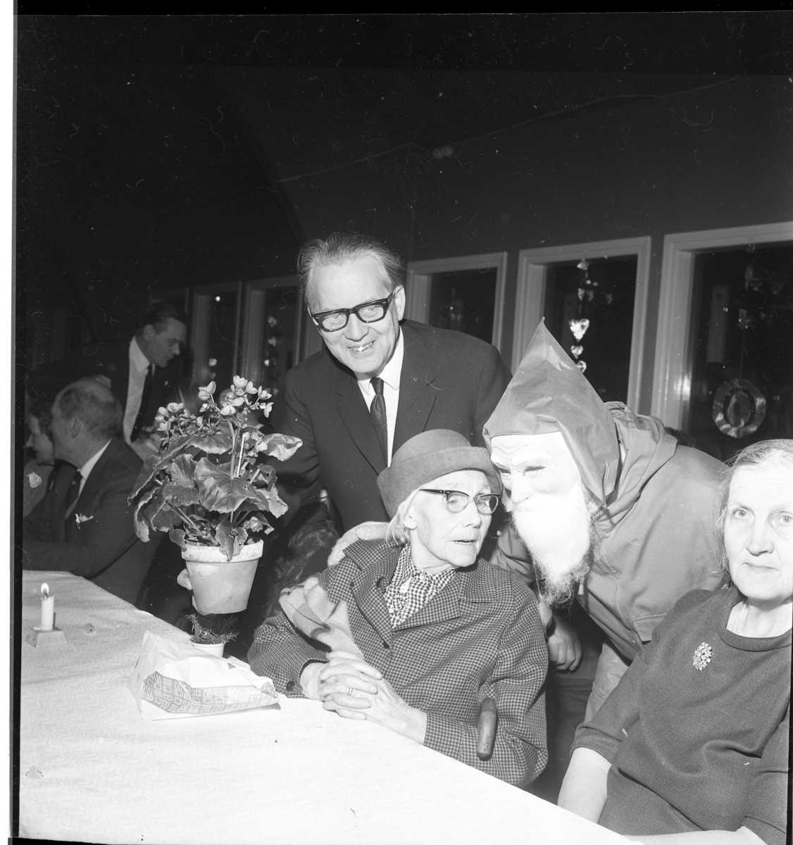 Män och kvinnor sitter vid långbord med ljus duk och tända ljus. En person med tomtemask böjer sig ned och vänder sig mot Hilma Samuelsson från Kleven som uppvaktas på 100- årsdagen. På hennes andra sidan lämnar Tage Grennfelt över en en blomma i en kruka.