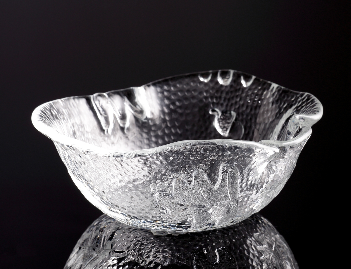 Asymmetriskt skål i ofärgat glas med reliefdekor av eklöv och ekollon.
Skålen är först gjuten till en "skiva" genom att man hällde ut glas över ett underlag med nämnda ekmönster. Sedan formades skålen i varmt tillstånd.
