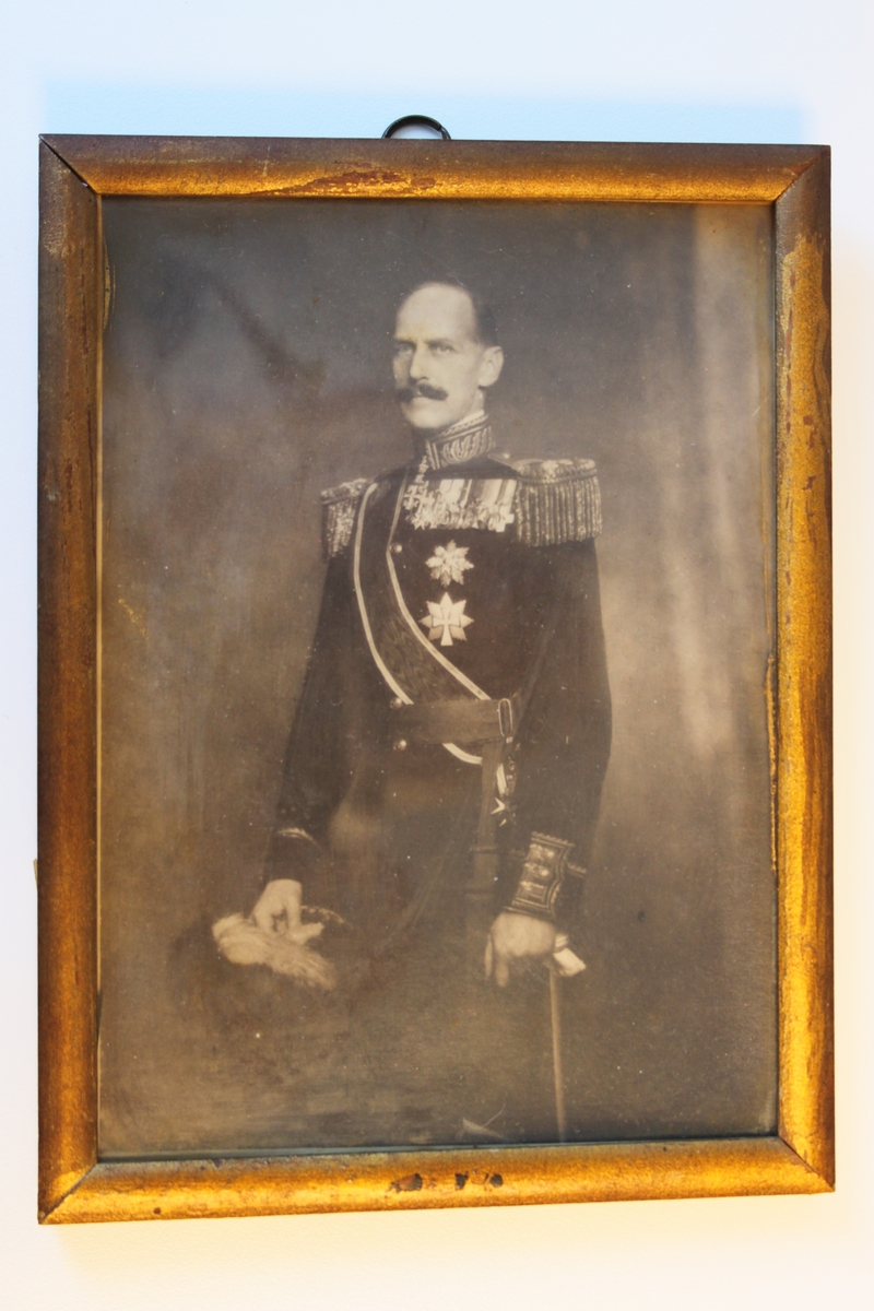 Bilde i Sort/hvitt. Portrett av Kong Haakon VII i Hærens gallauniform. Med gullramme i tre. Med oppheng i topp. Kanten på rammen i topp er vanlig skarp kant. I rammens bunn er kanten spikket litt rund inn mot baksiden.
