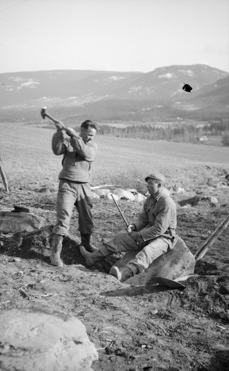 To mann med slegge og spett i gang med å dele en stor stein på jordet, antakelig på Linjordet i Østre Gausdal