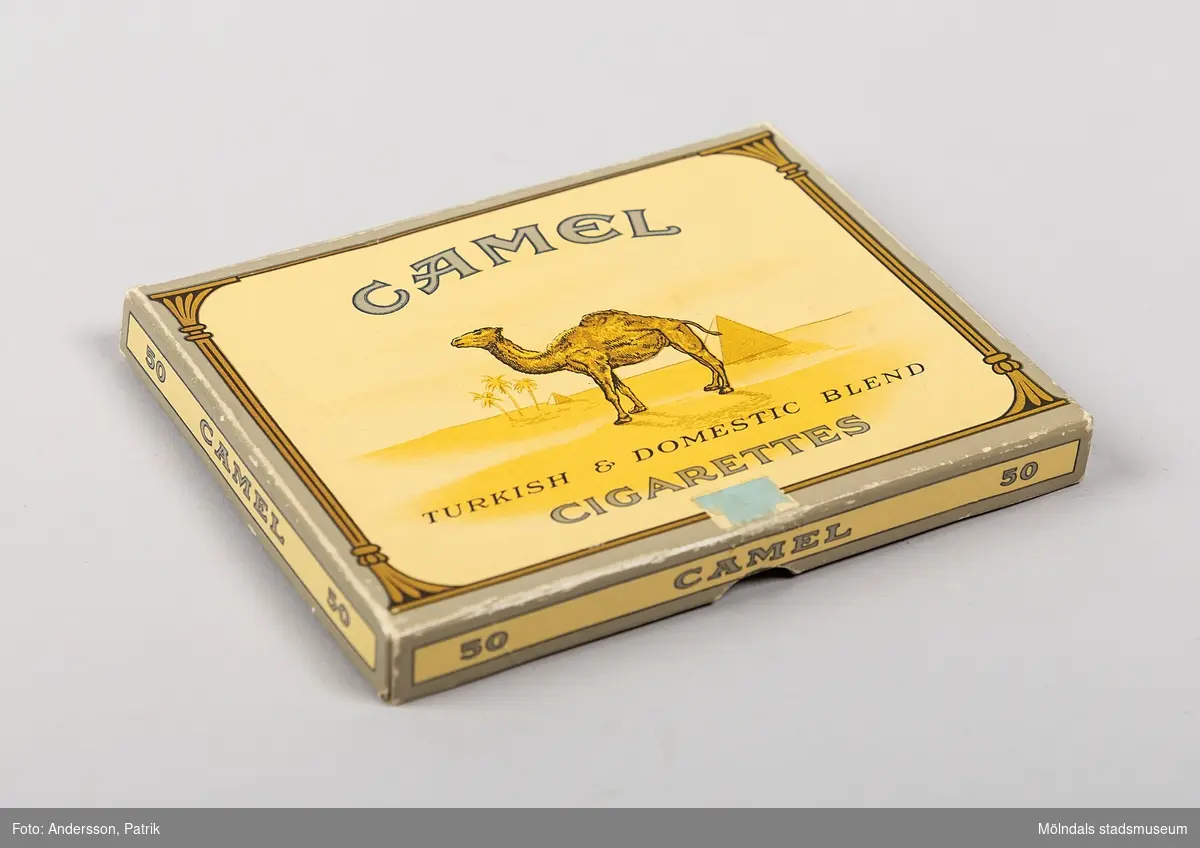 Pappask: Camel Turkish & Domestic Blend Cigarettes, som har innehållit 50 st cigaretter tillverkade av  R.J. Reynolds Tobacco Company troligtvis på 1950-talet.
Camel är ett amerikansk cigarettmärke som lanserades 1913. Deras cigarettpaket brukar vara kända för sin bild på en dromedar.

Cigarettsken är av papp och är gul. Längs kanterna är asken grå. På sidorna finns texten:  "50   CAMEL   50 ",  På askens ovansida finns texten:  "CAMEL TURKISH & DOMESTIC BLEND CIGARETTES", tillsammans med en dromedar. På insidan av locket finns texten: "CAMEL The Better CIGARETTE MANUFACTURED BY R..J. REYNOLDS TOBACCO COMPANY WINSTON - SALEM, N.C., U.S.A."
På undersidan av asken finns en varningstext ang. cigaretterna.