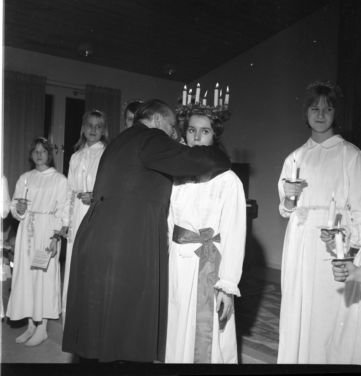 Ett luciatåg i Gränna står uppställda på rad. Kyrkoherde Wallis Wendel fäster ett halssmycke runt halsen på lucian Anna-Beth Petri. Två från vänster står Annika Andersson och till höger Margareta Olsson.