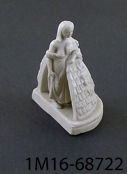 Leksak eller figurin av vit parian (porslin), föreställande dam. Ena halvan av damen är klädd, medan andra halvan av damen visar klädedräkten i genomskärning.