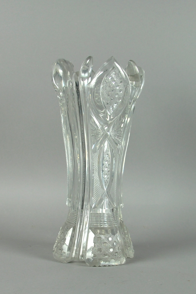 Vase av glass. Vasen er sylindrisk konkav, med innslipt dekor. Dekor består av ovale former med rombeformet kryssmønstre, rettslipte linjer og rutemønster.
