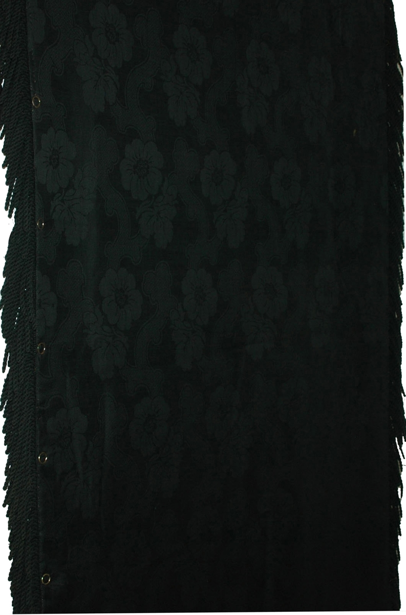Bordduk, svart korsstygnsbroderad botten med blommor i samma teknik i flera färger. Fodrad med svart, maskinvävt ylledamasttyg. Runt duken finns en drejad frans, ca 12 cm.
Försedd med ett antal metallöljetter av mässing på baksidan.