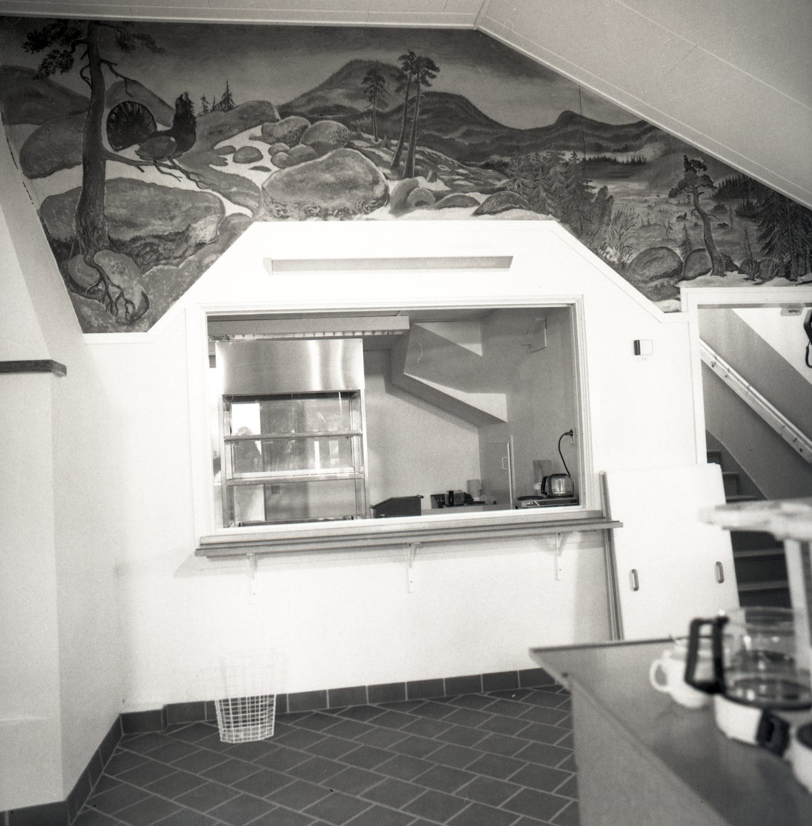 Ett rum med ett litet kök med servering, det finns en monter för bakverk och kaffepannor och kaffemuggar finns det. Ovanför den lilla serveringsluckan är det en väggmålning och man ser en trappa. 1992 Grytan Brunflo.