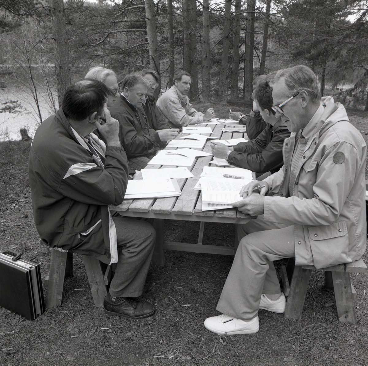 På några bänkar vid ett bord sitter det en grupp män och bläddrar i sina papper, bredvid på marken står en portfölj.  Nedanför finns det träd, vatten och skog. Länsyrkesrådet och Skogsbruksskolan Törn 5 juni 1990.
