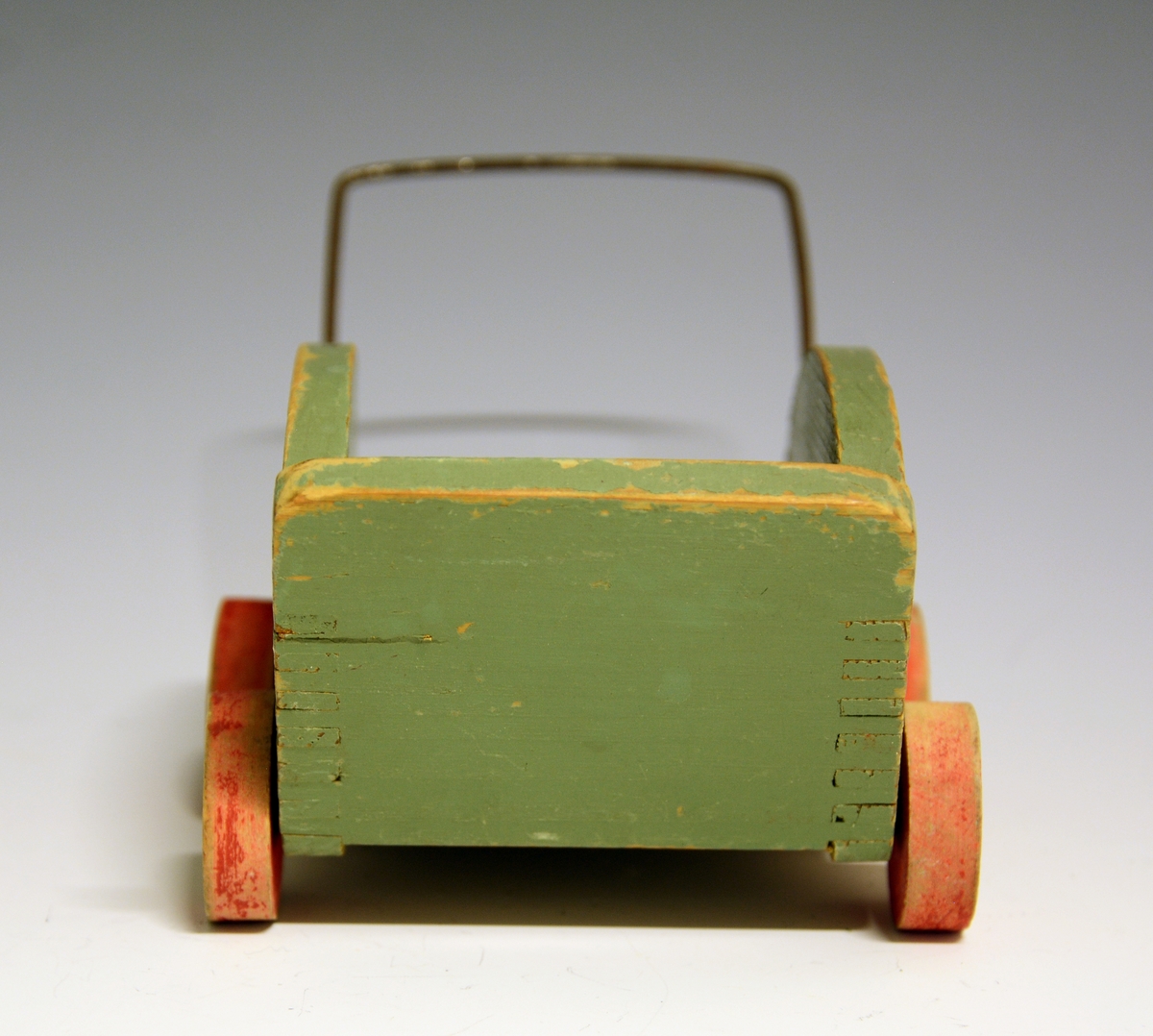 Hjemmelaget dukkevogn i tre med fire hjul og håndtak av metall. Grønnmalt, borsett fra hjulene som er røde.