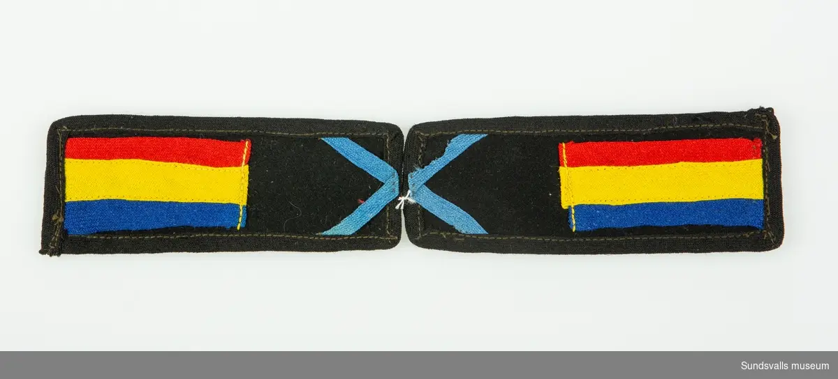 Axelklaffar i svart ylle. Blå vinklade band i ena änden, blå-, gul- och rödrandigt band i den andra änden på båda klaffarna.