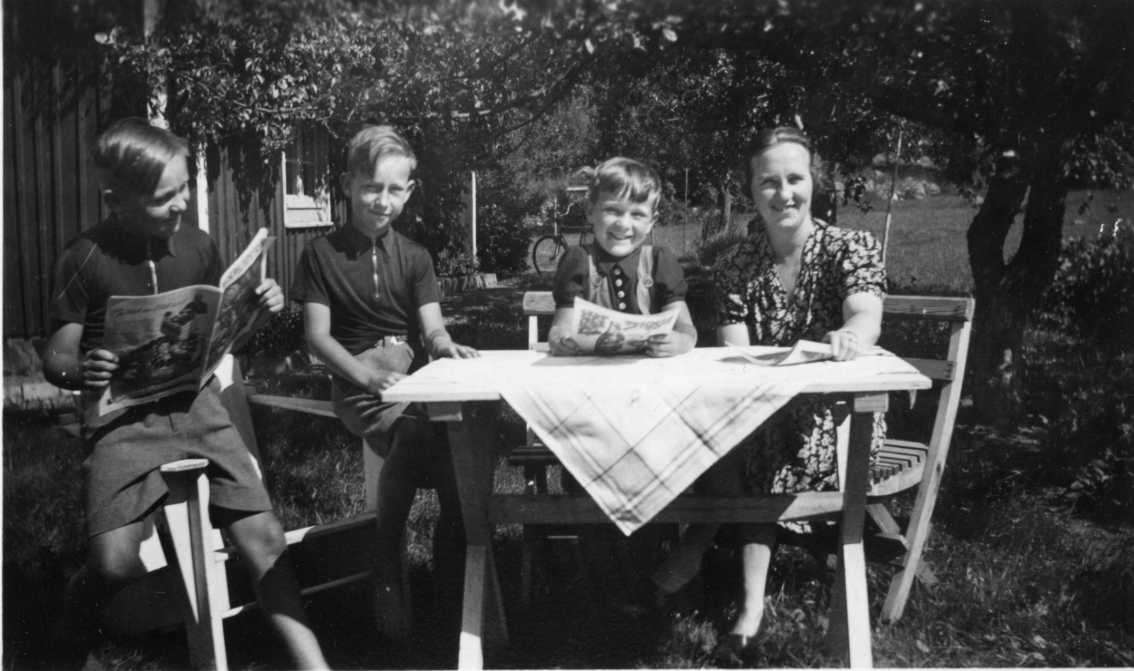 Tre pojkar i kortbyxor och kortärmad tröja sitter ute vid ett trädgårdsbord med sin mamma, Nanna, till höger i blommig klänning. Pojkarnas namn är Åke, Ingemar och P-G. Pojken till vänster tittar i tidningen Husmodern. Pojken till höger liksom mamman håller i varsin tidning. De skrattar och ler mot fotografen. Det är sommar.