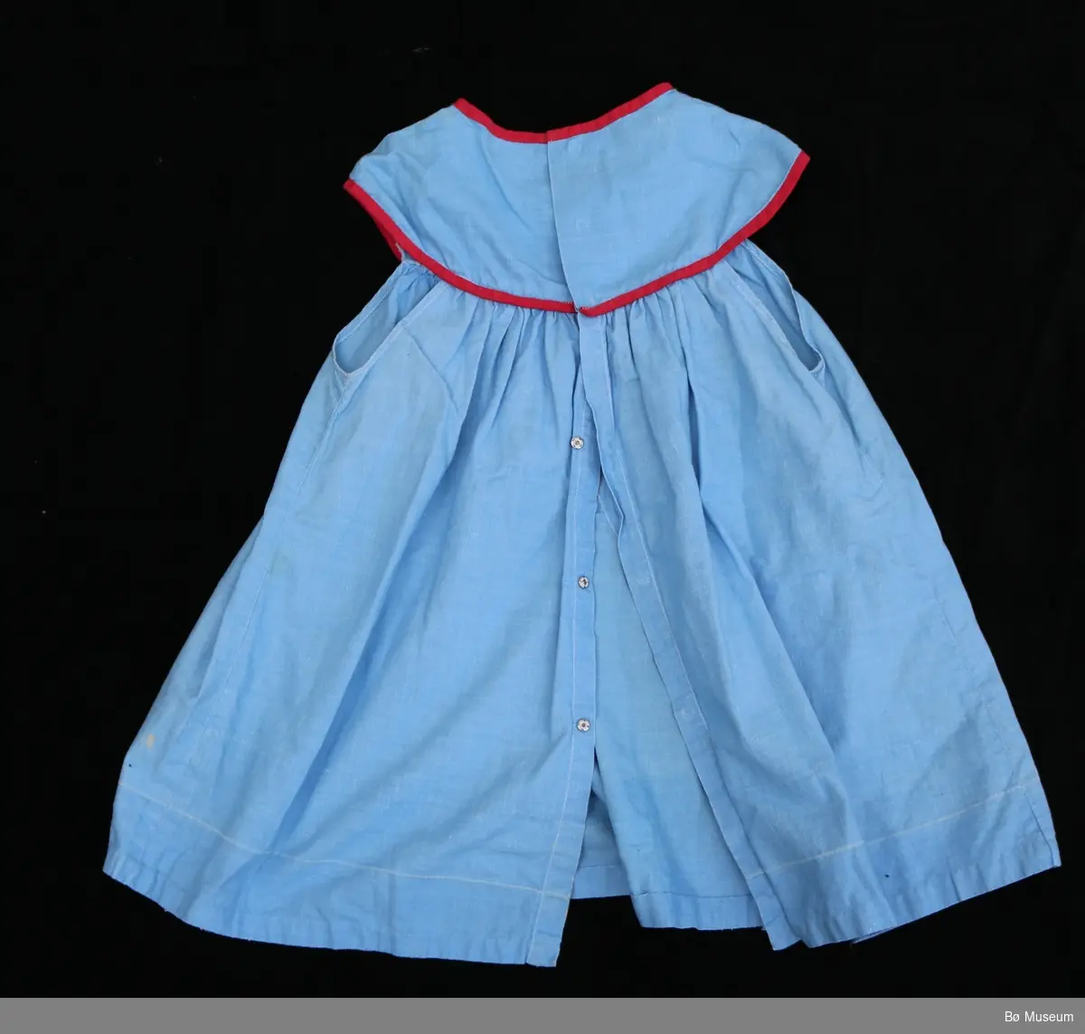 Blå babykjole med raud kanting.
Foran er er to små lommer.