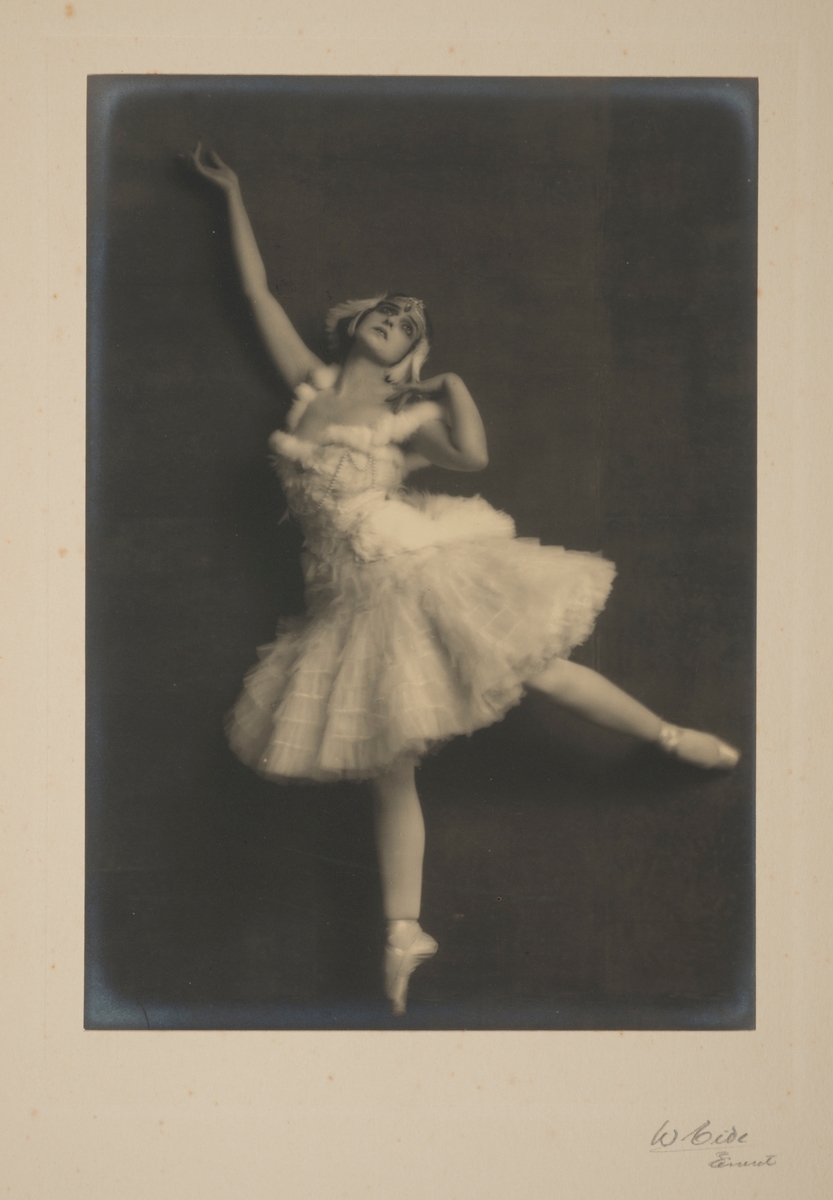 Kunstnerisk portrett av den berømte russiske ballettdanseren Vera Fokina, mens hun danser (tåpiss) i hvit ballettkjole.
Fra 'Svanens død'.